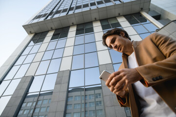 Fototapeta premium Bottom view of good looking businessman in autumn coat using cellphone near business center 