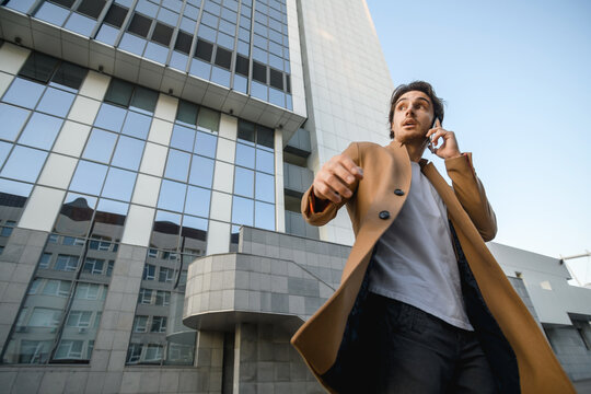 Low angle view of businessman in coat talking on mobile phone during wind near skyscraper outdoors 