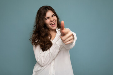 Portrait of young positive happy joyful attractive curly brunette woman with sincere emotions wearing casual white shirt isolated on blue background with empty space and pointing at camera