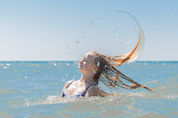 Young attractive girl teenager of European appearance throws wet, long hair up into the sea against the background of the horizon line and blue sky