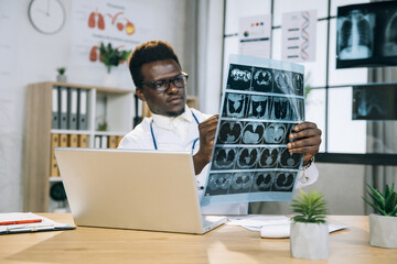 Focused african doctor in eyeglasses analysing tomography while sitting at modern office. Competent medical specialist using laptop while making diagnosis for patient. Selective focus on xray scan