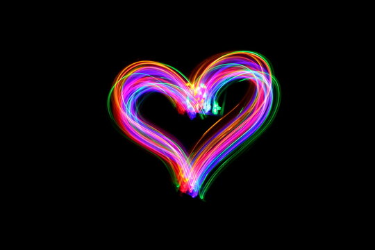 Long exposure photograph of a heart shape outline in neon colour in an abstract swirl, parallel lines pattern against a black background. Light painting photography.