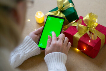 A phone with a green screen in the hands of a woman in a white sweater, a red and green gift box and bokeh lights on the table. Close up. Concept of online shopping for Christmas and New Year