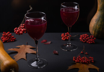A crimson drink in a glass with a bunch of mountain ash, around autumn leaves, oblong pumpkins, mountain ash on a gloomy dark background. Autumn still life with halloween mood.