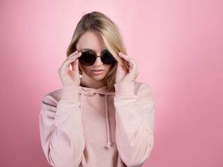 beautiful young woman with blonde hair wearing dark sunglasses looking into the camera. In a pink hoodie on a pink background, studio portrait