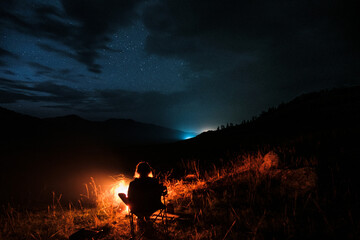 A man sits by a fire at night in the mountains against a backdrop of stars