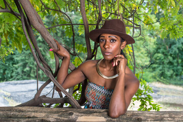 Wearing a brown outback hat,a golden neck ring, and one arm supporting her head, a pretty black girl is standing outside in a small woods and looking at you..
