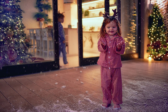 Happy Toddler Baby Girl Catching Snowflakes At The Patio On Christmas Night. Kids Having Fun At Home During Winter Holidays