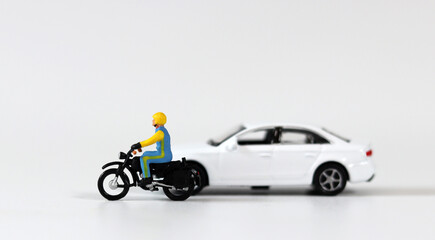 A miniature driver wearing a helmet next to a white miniature car and riding a motorcycle....