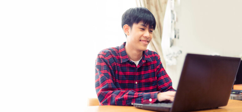 Happy Young Asian Male Smiling Using Laptop At Home.Work From Home.Isolated Banner Background.Teenager Young Boy Online Learning Typing Social Chat.Social Media.University Male, Blogger Freelance Man.