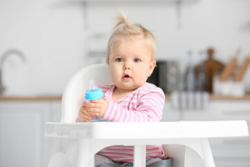 Cute baby girl with bottle of water sitting on high chair in kitchen