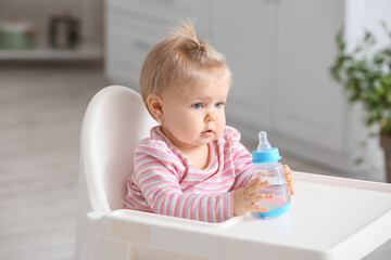 Cute baby girl with bottle of water sitting on high chair in kitchen