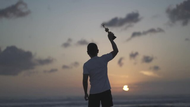 Silhouette of a proud man lifting a trophy on the beach at sunset - Winner Achievement in Slow Motion