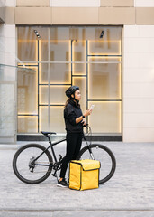 Side view female messenger standing outdoors with bicycle and thermal backpack holding a smartphone looking for a customer address