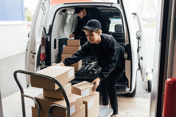 Two people in uniform loads parcels in van. Woman taking boxes from cart.
