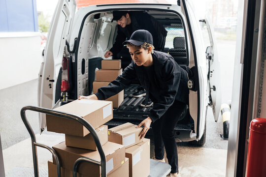 Two people in uniform loads parcels in van. Woman taking boxes from cart.