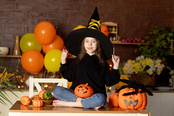 halloween, child a girl in a witch costume and a hat in a dark kitchen with pumpkins and a big spider is fooling around, smiling, rejoicing