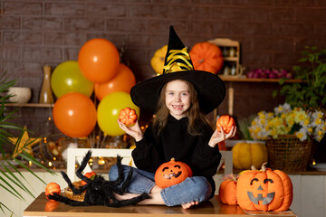 halloween, child a girl in a witch costume and a hat in a dark kitchen with pumpkins and a big spider is fooling around, smiling, rejoicing