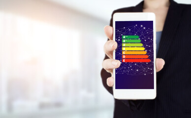 Good energy chart rating. Hand hold white smartphone with digital hologram energy efficiency sign on light blurred background. Energy efficiency concept.
