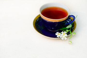 A cup of tea and a small bouquet of white flowers on a white table, a close - up view. Black or herbal morning or afternoon tea in the open air. Hot tea in a beautiful blue cup with a gold rim