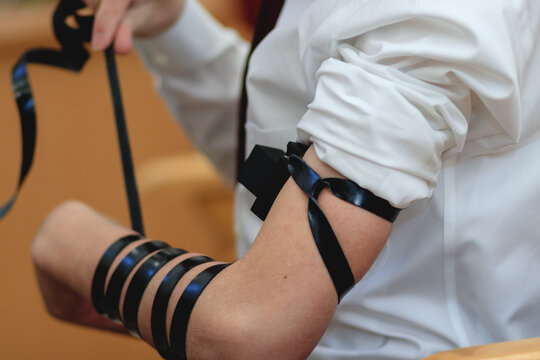  A close-up of a Jewish bar mitzvah boy, wearing a white shirt, put s tefillin for the first time in a synagogue