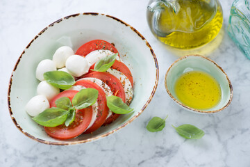 Caprese salad with mozzarella, red tomatoes and green basil leaves served in a green bowl, horizontal shot on a light-grey marble background