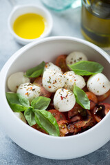 Close-up of caprese salad with mini mozzarella balls, red cherry tomatoes and fresh green basil served in a white bowl, vertical shot, selective focus