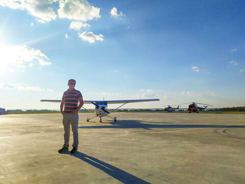 A young, handsome male pilot in casual clothes, glasses standing near a small pleasure plane on the airfield. Aviator stands on a runway near an aircraft on a landing point with private jet terminal.