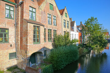 Fototapeta na wymiar Brugge, Belgium - October 9. 2021: View along water canal with green trees and old residential brick house against clear blue sky