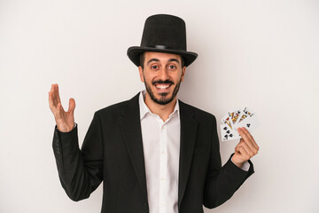 Young magician man holding a magic card isolated on white background receiving a pleasant surprise, excited and raising hands.