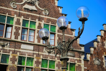Gent, Belgium - October 9. 2021: Closeup of three armed old street lamp with typical belgian stepped gable house facade background against blue sky