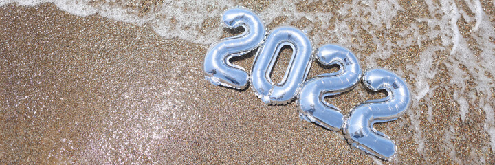Silver balloons with numbers 2022 lying on sand by sea closeup background