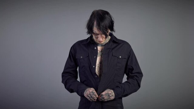 Portrait of young man with tattooed torso buttoning black shirt looking at camera with confident facial expression. Caucasian brunette guy posing at grey background dressing up
