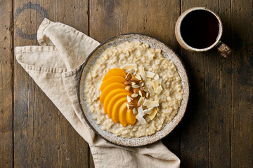 Bunting porridge with sliced mango, chopped almond nuts, coconut chips and cup of tea. Traditional oatmeal breakfast served in ceramic bowl on wooden background