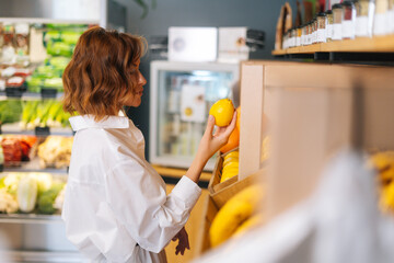 Side view of attractive young woman choosing fruits for diet, checking lemon in grocery store at...