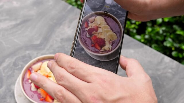 Male hands making photo, picture, shots for social media of healthy fruit and berries smoothie bowl on smartphone. Man, influencer, blogger shoot on phone camera breakfast, restaurant food. Close-up.