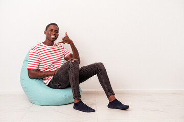 Fototapeta premium Young African American man sitting on a puff isolated on white background showing a mobile phone call gesture with fingers.
