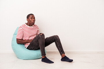 Fototapeta premium Young African American man sitting on a puff isolated on white background shrugs shoulders and open eyes confused.