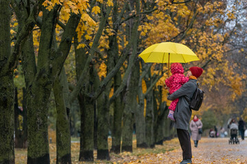 Mom holds a small child in her arms while standing under a yellow umbrella in an autumn park against a background of yellow foliage