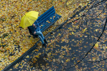 Top view. Man sits on bench in the yard under yellow umbrella, surrounded by fallen leaves.