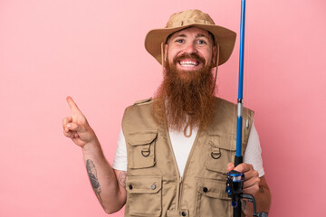 Young caucasian ginger fisherman with long beard holding a rod isolated on pink background smiling and pointing aside, showing something at blank space.