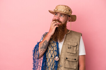 Young caucasian ginger fisherman with long beard holding a net isolated on pink background