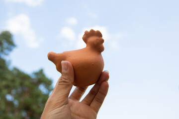 A female hand holds a clay cockerel against the background of the sky. Folk craft. Cock-shaped whistle