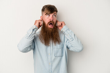 Young caucasian ginger man with long beard isolated on white background covering ears with fingers,...