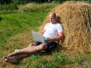 Naklejka na ściany i meble a middle-aged man, dressed in a T-shirt and shorts, is lying on a yellow haystack with a gray laptop in his hands. Remote work, training. Rural area. Thoughtful facial expression, side view
