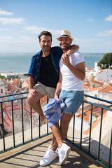 Smiling gay couple on sunny day. Two men in casual clothes, standing on terrace balcony, sea in...