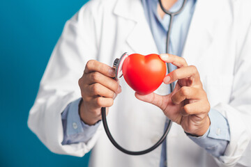 Hand of a doctor holding a stethoscope checking a red heart shape while standing in the hospital or clinic. Close-up photo. Space for text. Medical, treatment, and healthcare concept