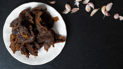 Top view dark tone of Fried sliced pork with garlic cutlets are placed on a white plate. on a black tiled floor with decorative garlic. Space to fill in the text.