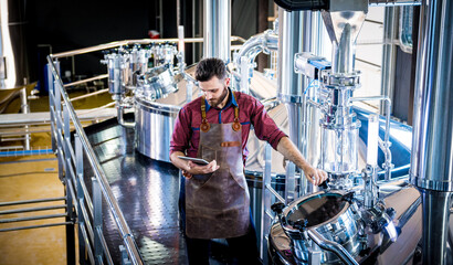 Young male brewer in leather apron supervising the process of beer fermentation at modern brewery factory - Powered by Adobe