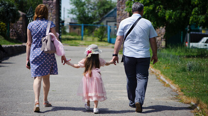 Obraz premium family walking in the park. mom dad and little girl walking holding hands. back view. daughter in a pink dress. friendly loving family concept, love. outdoor, walk in the fresh air, threesome